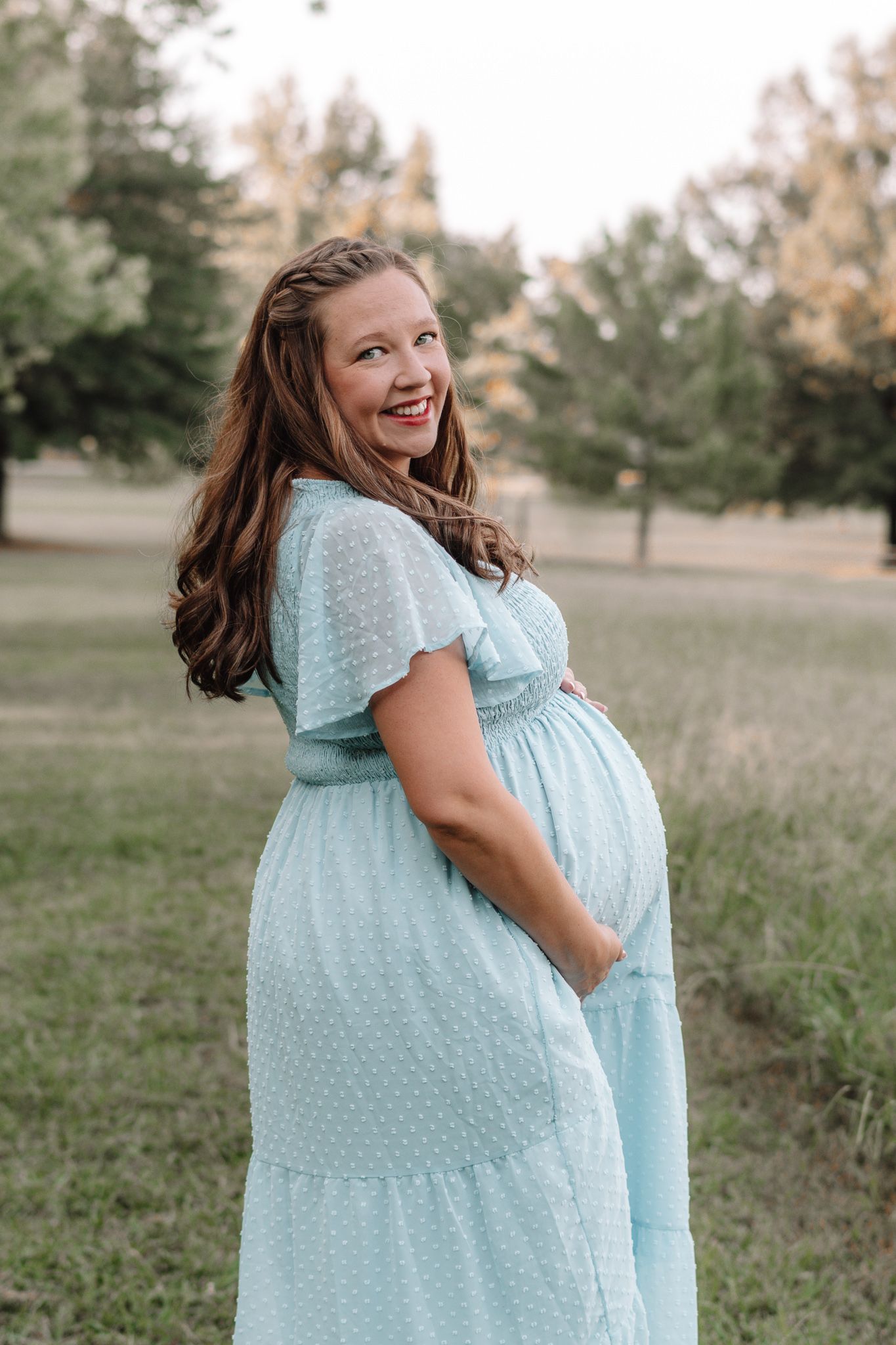 Joyful poses in a light blue dotted dress during an outdoor maternity photoshoot.