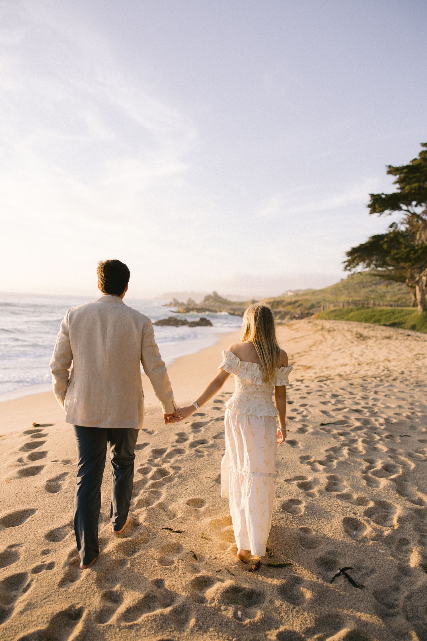 A couple walks hand in hand on a sandy beach at sunset with waves and coastal rocks in the background.
