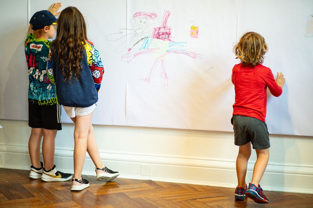 Three students draw with markers on a white wall in a studio or classroom space during an activity.