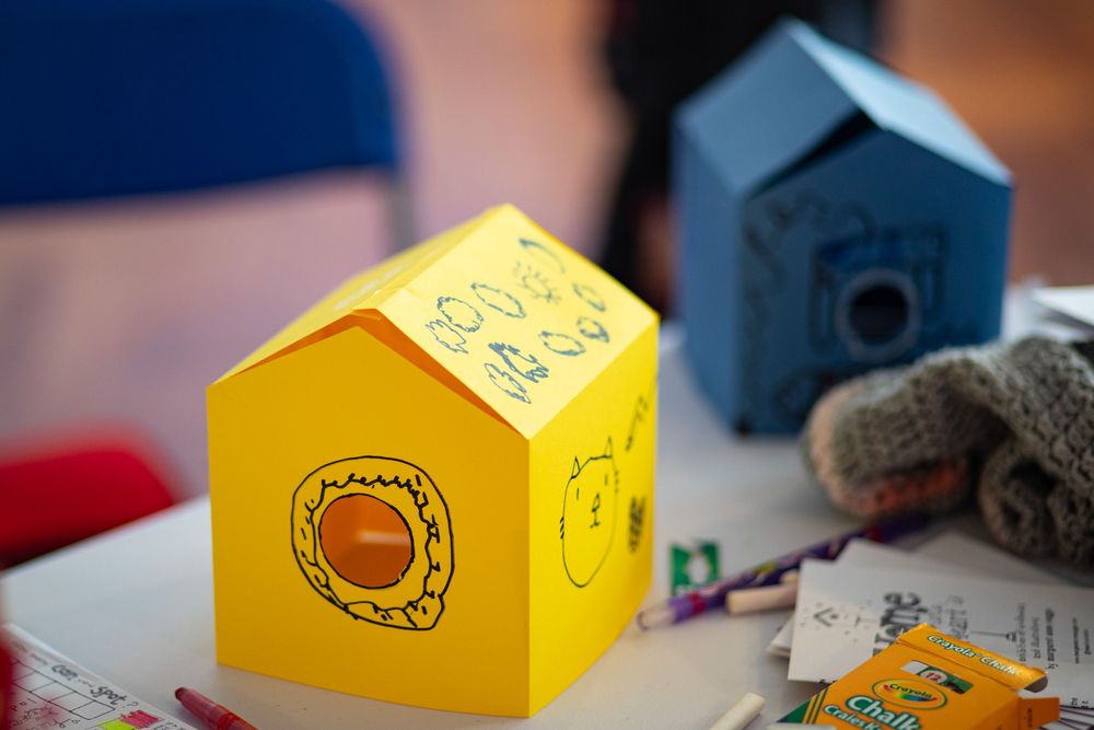 Small yellow house-shaped box with a decorative circular design sits on a table with a blue house and plush toy visible.