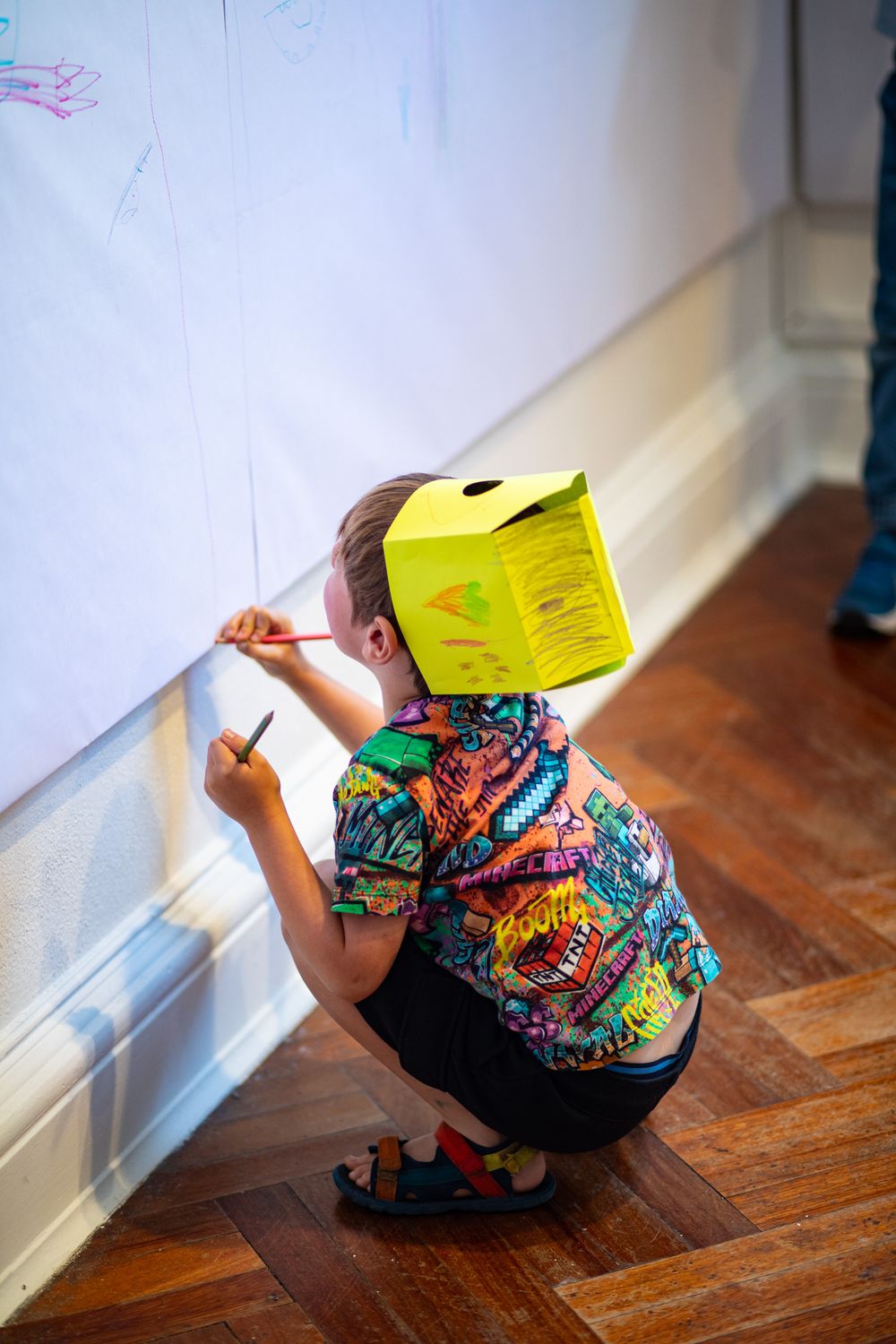 Person wearing colorful comic-print shirt crouches to write on wall with yellow box on head in indoor setting.