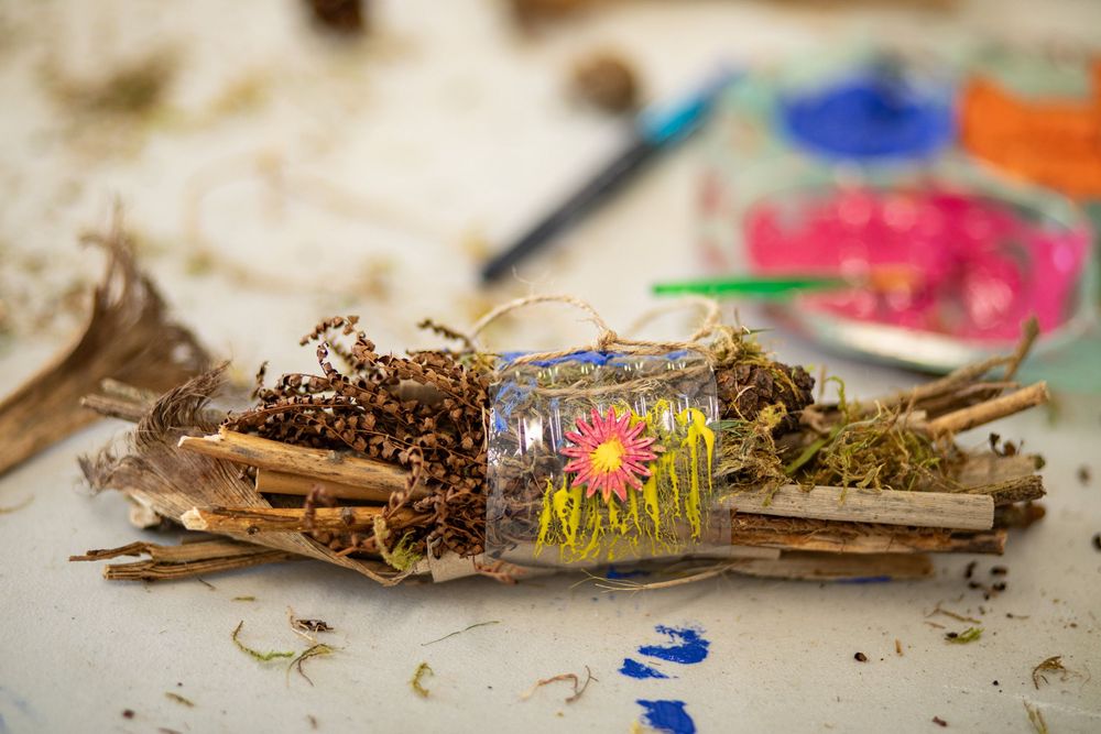 A small glass jar filled with a colorful artificial flower, nestled among twigs and dried foliage.