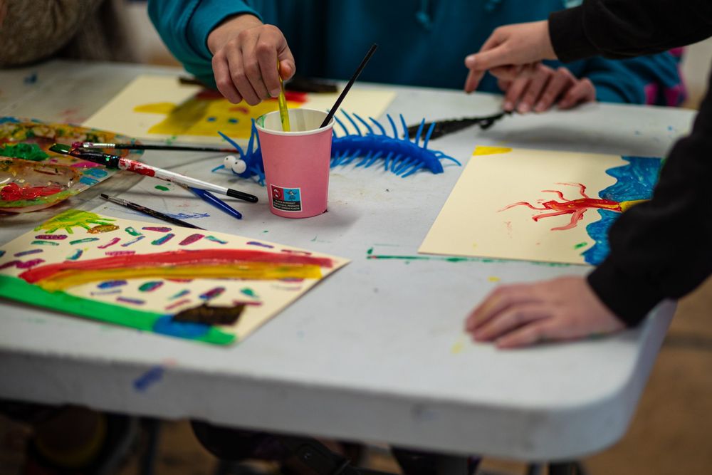 Children's hands visible around a table with colorful artwork, paint supplies and a pink cup with blue brush.