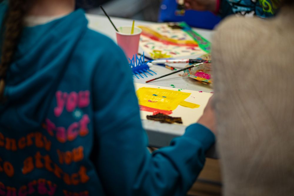 Someone wearing a blue sweater sits at a table with colorful art supplies and paper materials spread out.