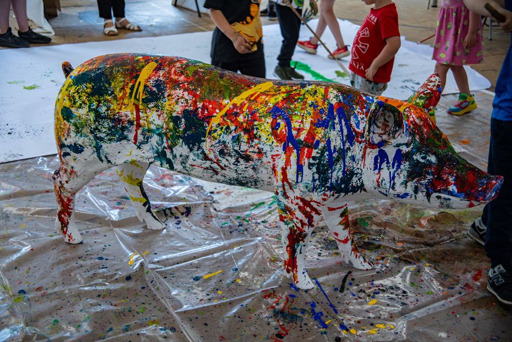 A multicolored sculpture of a cow covered in dripping paint stands on a messy art-covered floor at an art event.