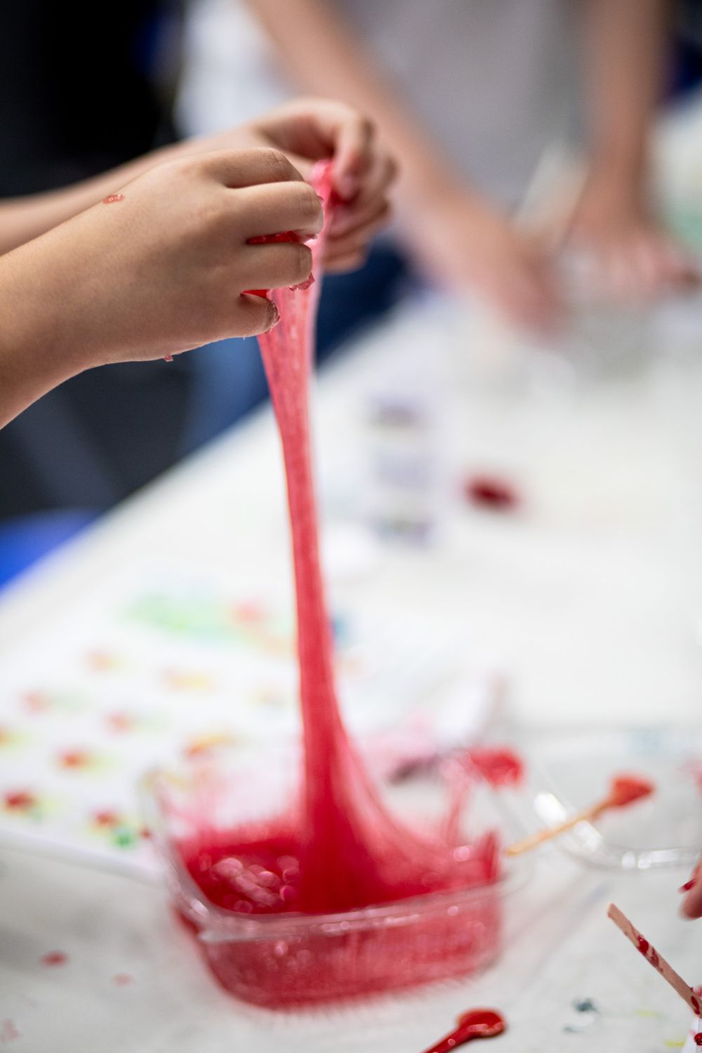 Close-up of hands playing with bright red slime or stretchy putty on a white surface during a sensory activity.