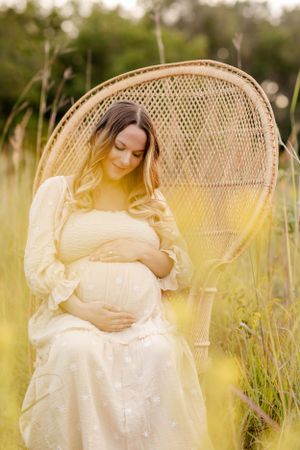 A pregnant woman in a white dress sits peacefully in a wicker peacock chair amid golden sunlit wildflowers.