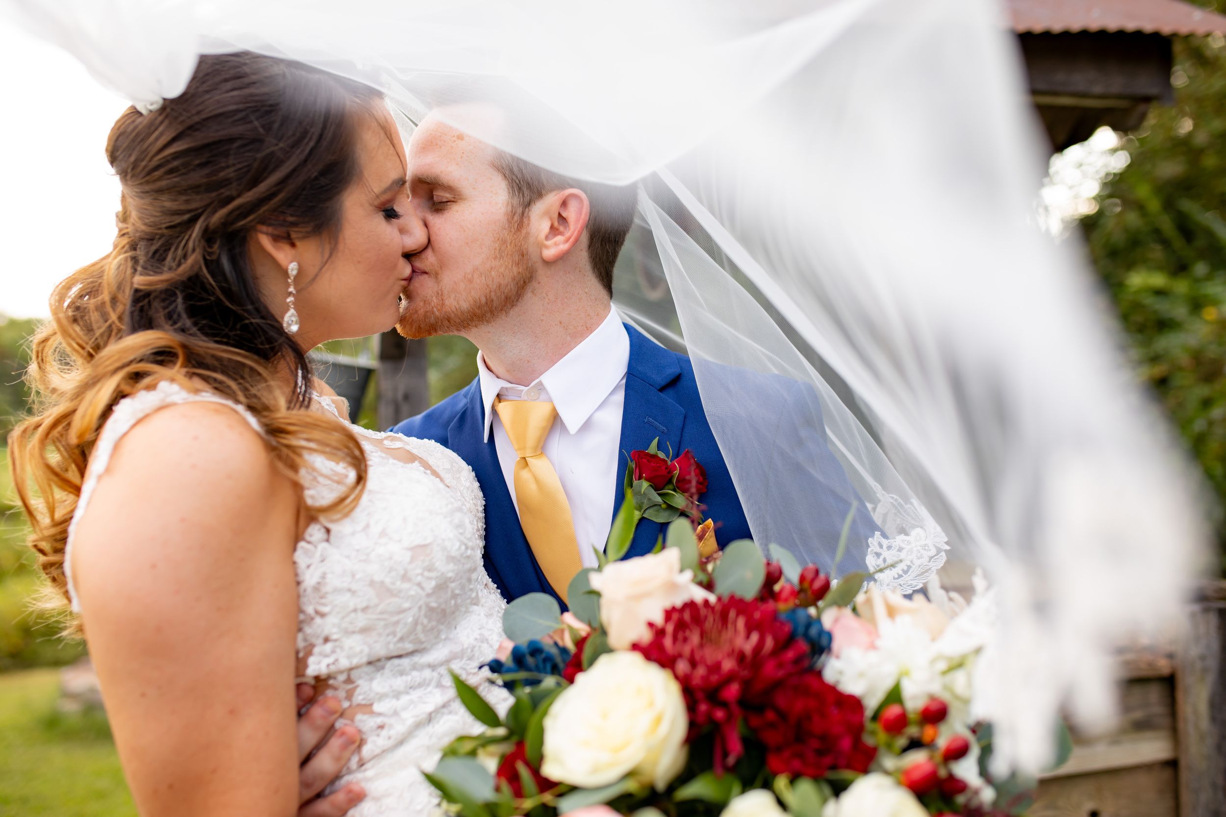 A newlywed couple shares a romantic kiss under a flowing veil while holding a red and white rose bouquet.