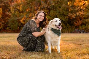 A golden retriever wearing a bandana poses with someone in a floral dress on autumn grass with colorful foliage behind them.