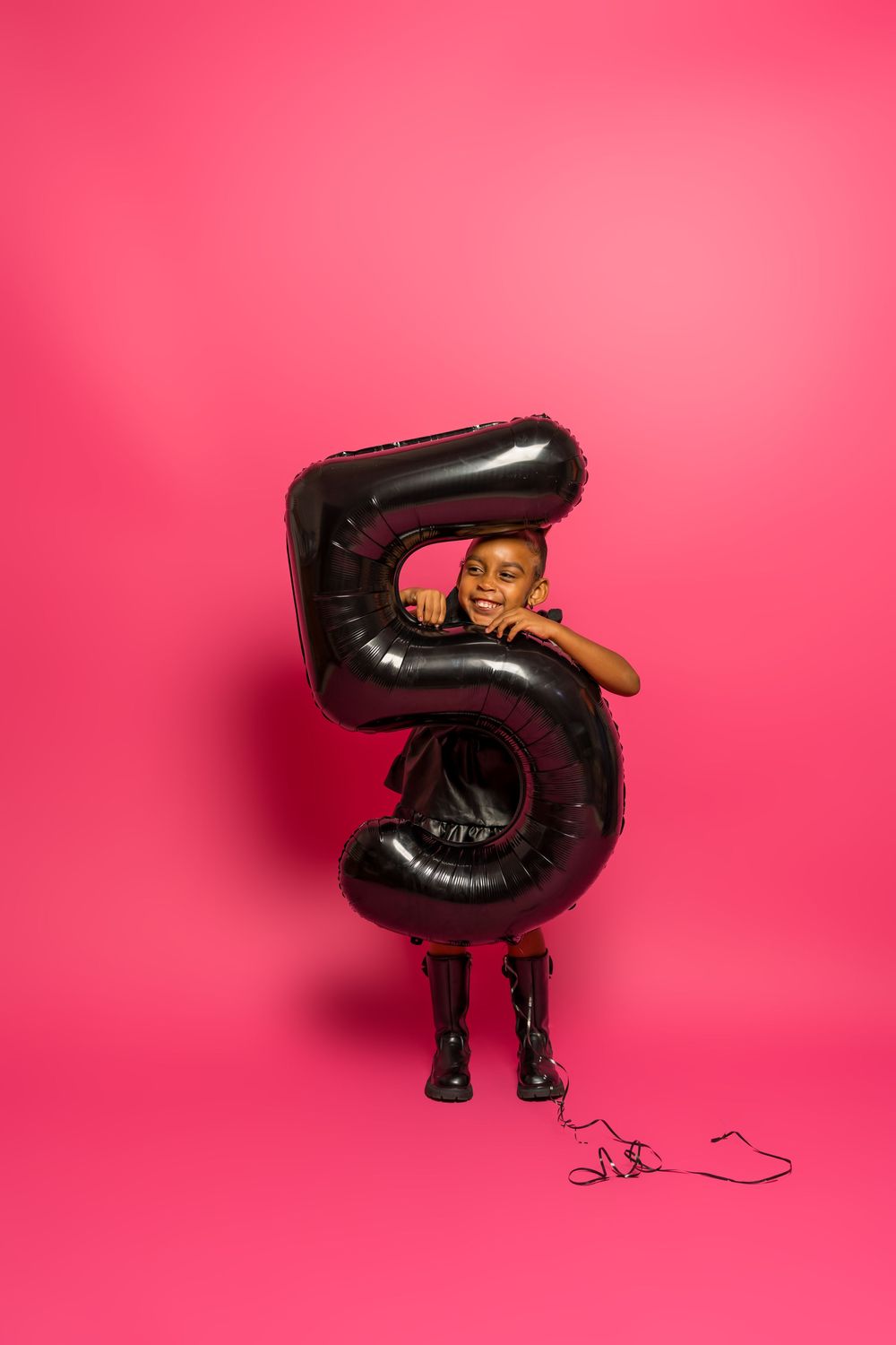 Child in black dress hugs large number 5 balloon against vibrant pink background.