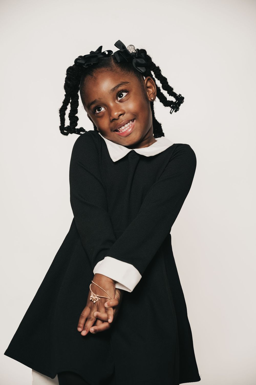 Child in classic black dress with white collar poses elegantly against white background.