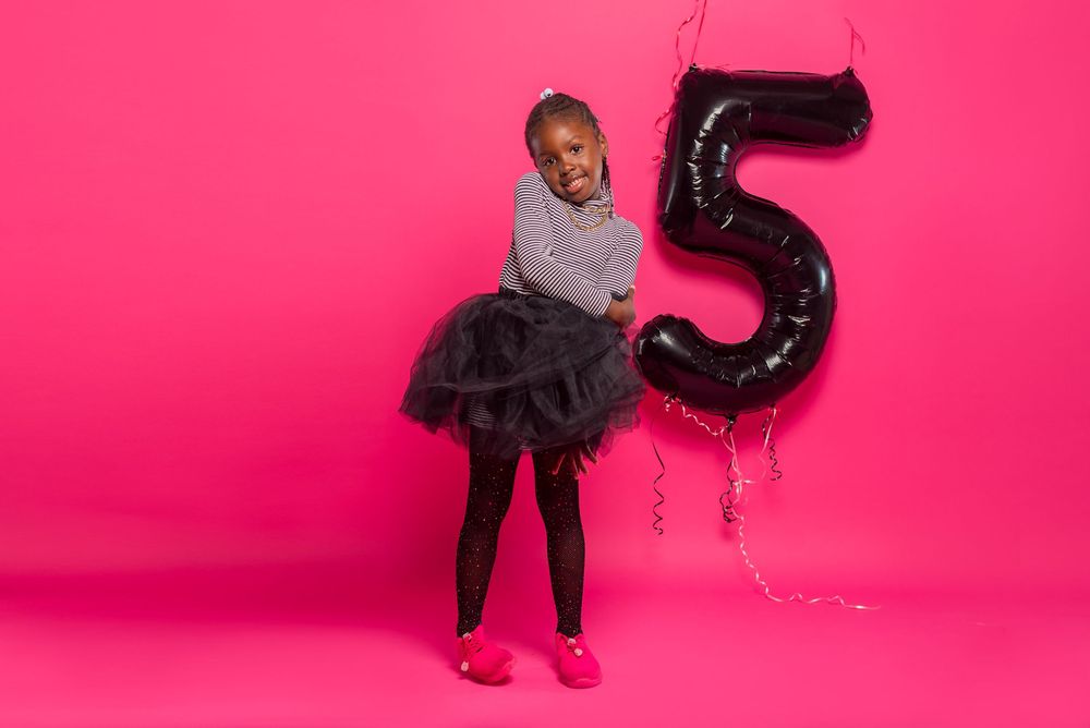 Child in tulle skirt poses with large black number 5 balloon on pink backdrop.