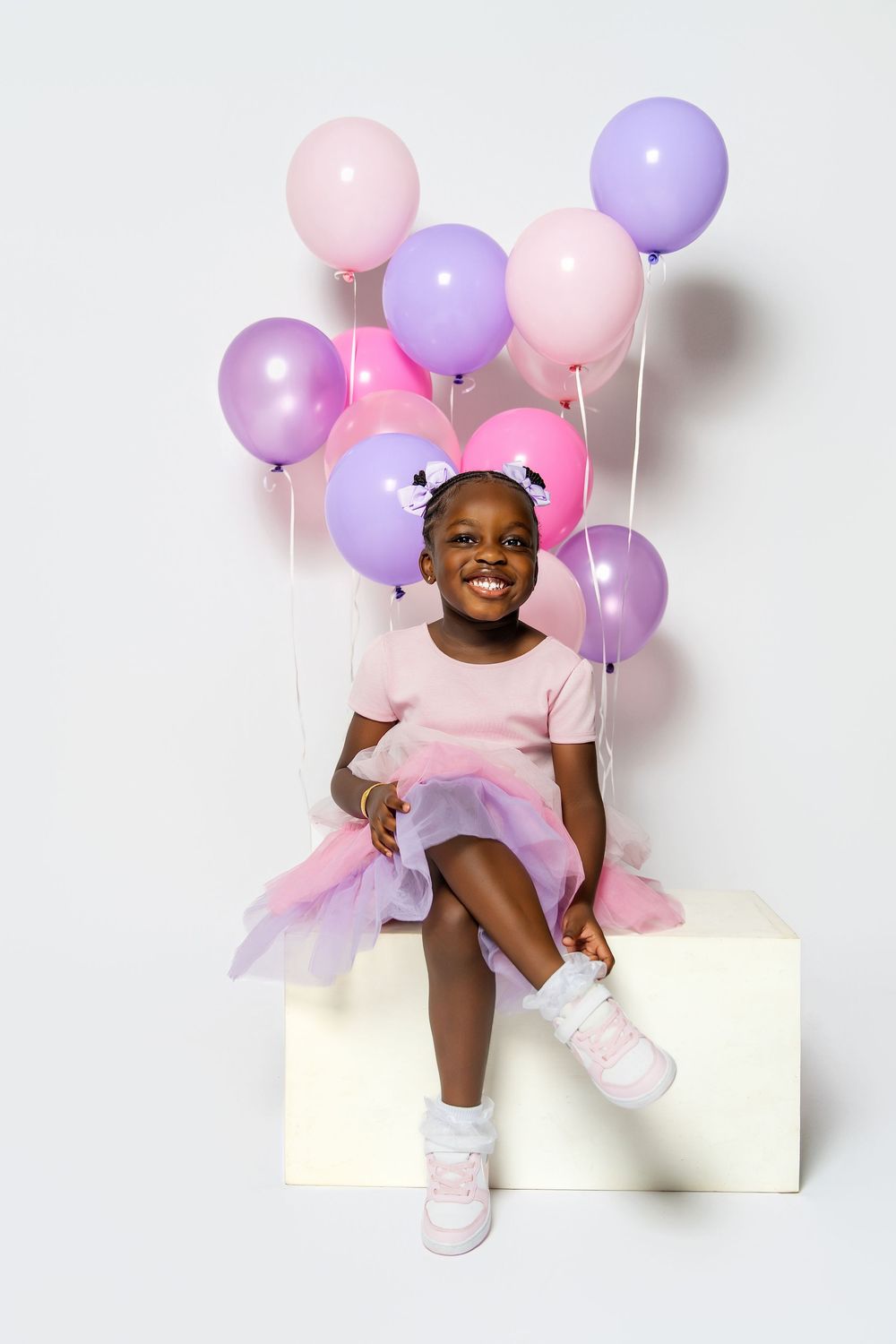 A young child in pink sits on a white platform surrounded by pink and purple balloons for a birthday photoshoot.