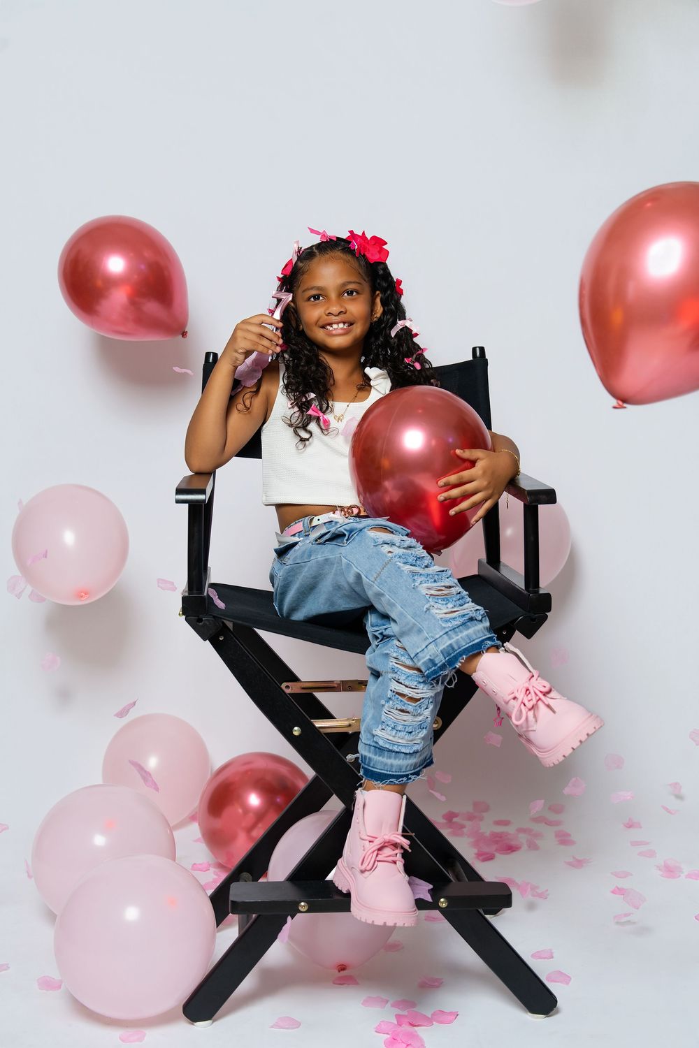 Young girl in ripped jeans poses playfully with pink and red balloons on a director's chair.