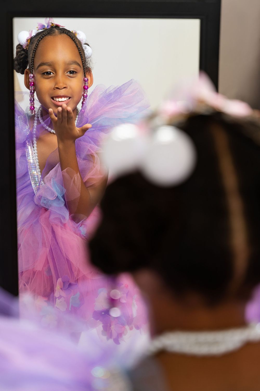 Young dancer with beaded braids in purple dress smiles at reflection in mirror.