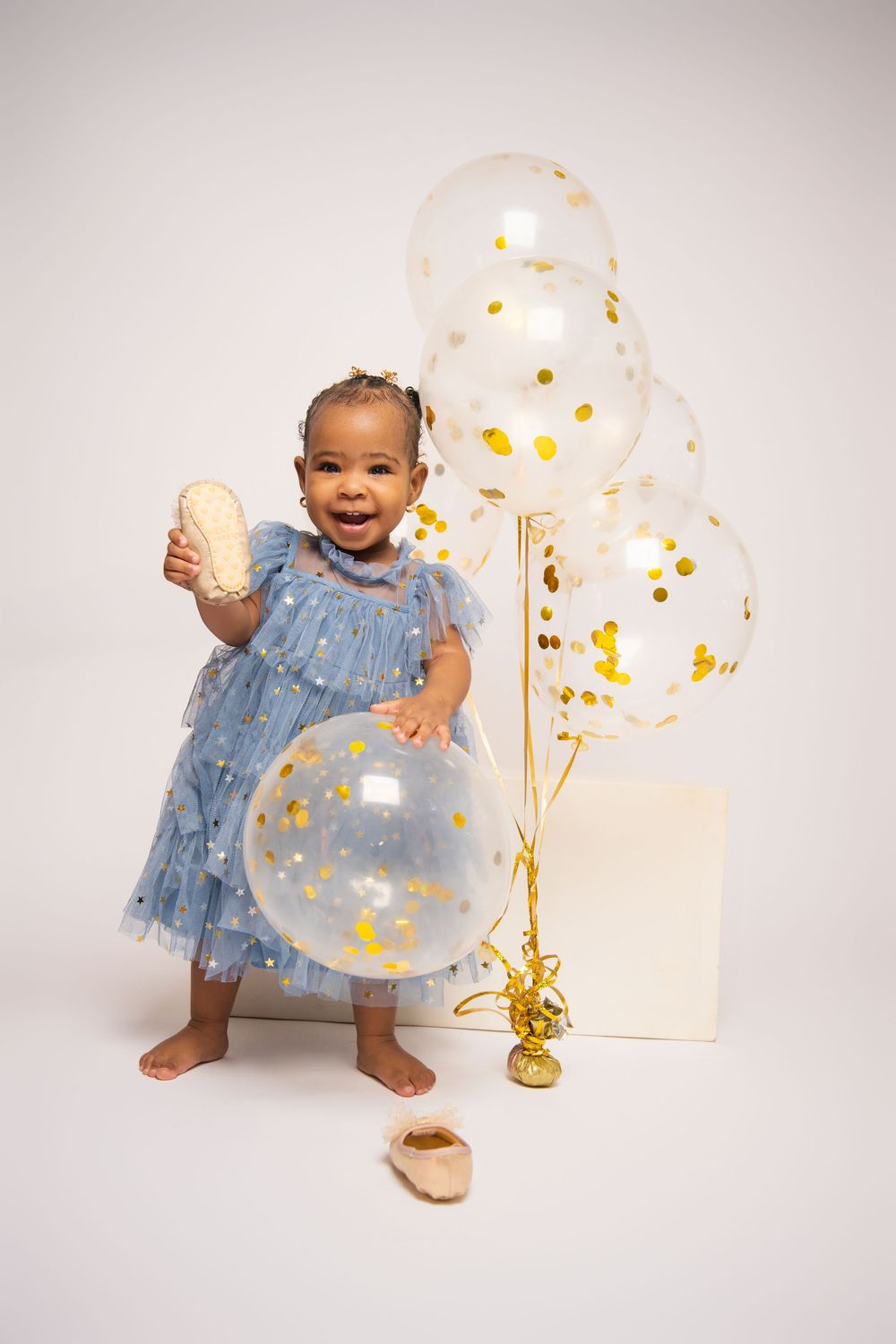 A happy toddler in a blue dress plays with gold confetti balloons in a studio setting.