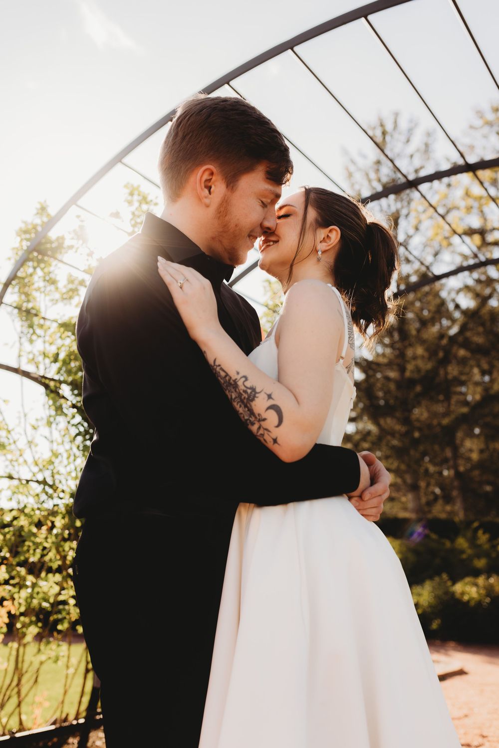 Intimate wedding portrait of couple sharing tender moment under garden arch at sunset.