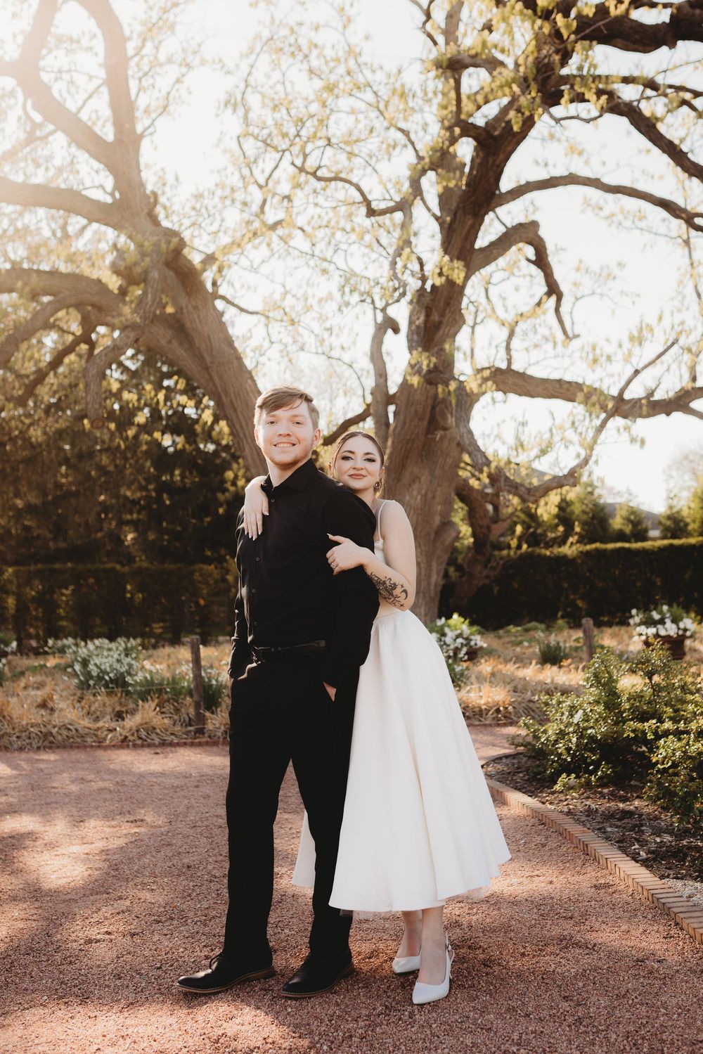 A couple in wedding attire poses beneath sprawling tree branches on a garden path.
