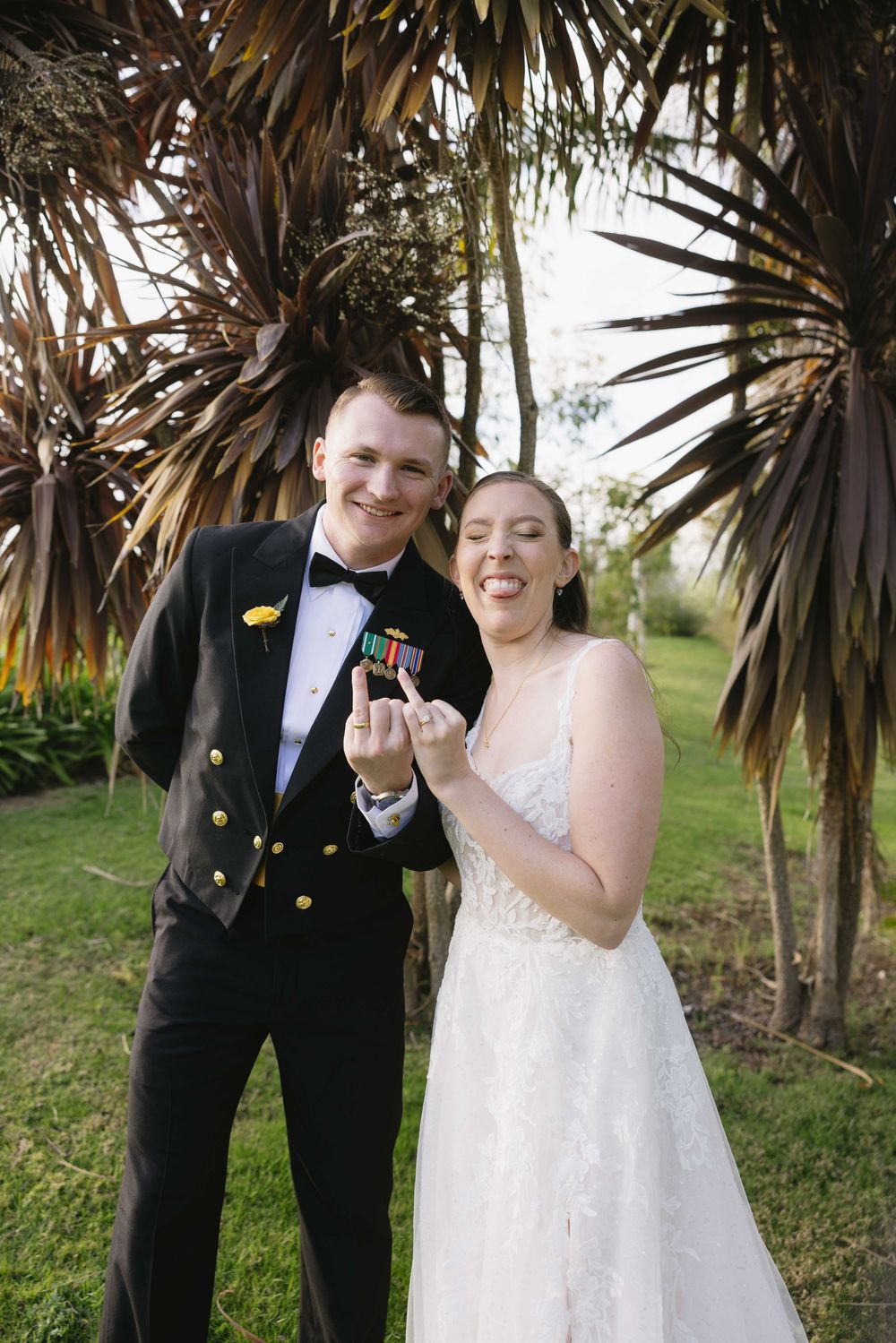 A joyful wedding couple poses outdoors beneath palm trees while holding a small prop.