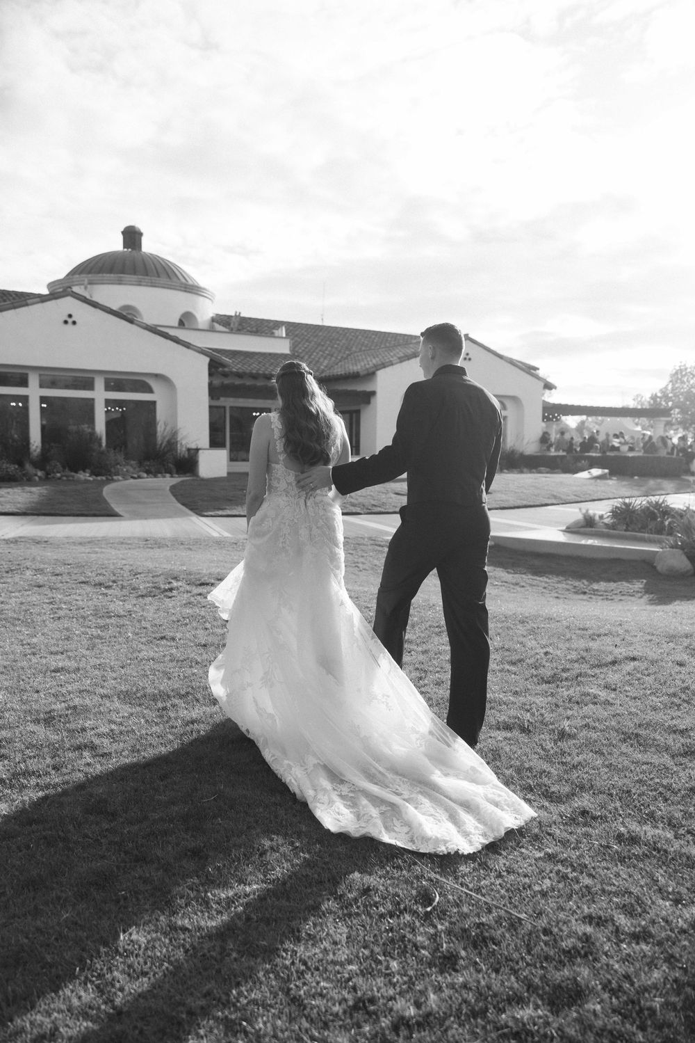 A romantic black and white photo of newlyweds walking on grass with a Spanish-style building in the background.