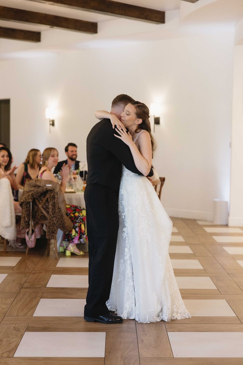 Newlyweds share their first dance on a checkered tile floor as wedding guests applaud in the background at a reception.