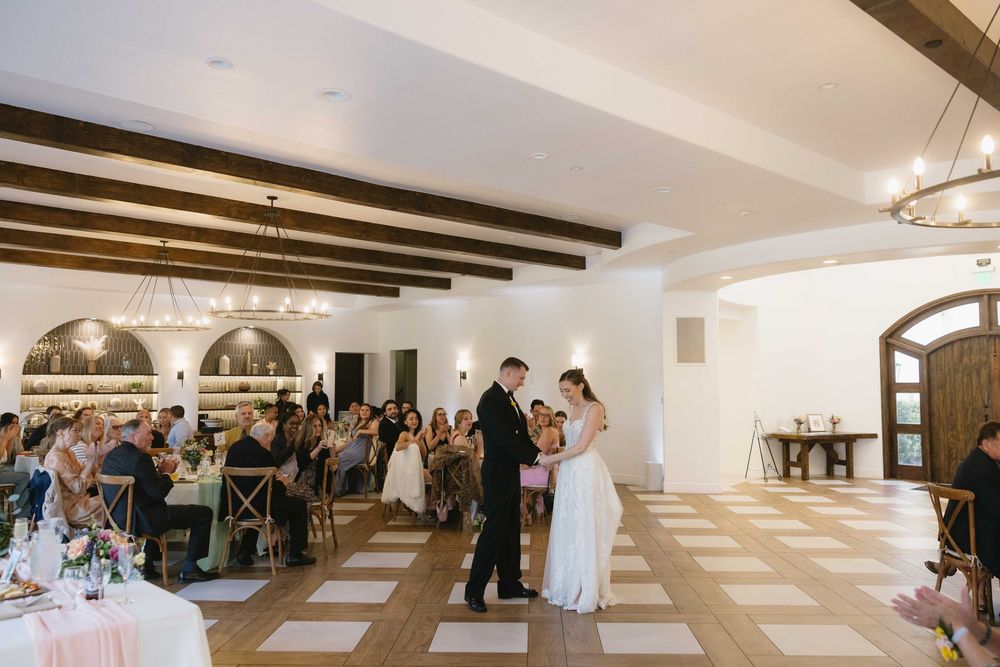 Couple shares their first dance at an elegant reception venue with exposed wooden beams and arched windows.