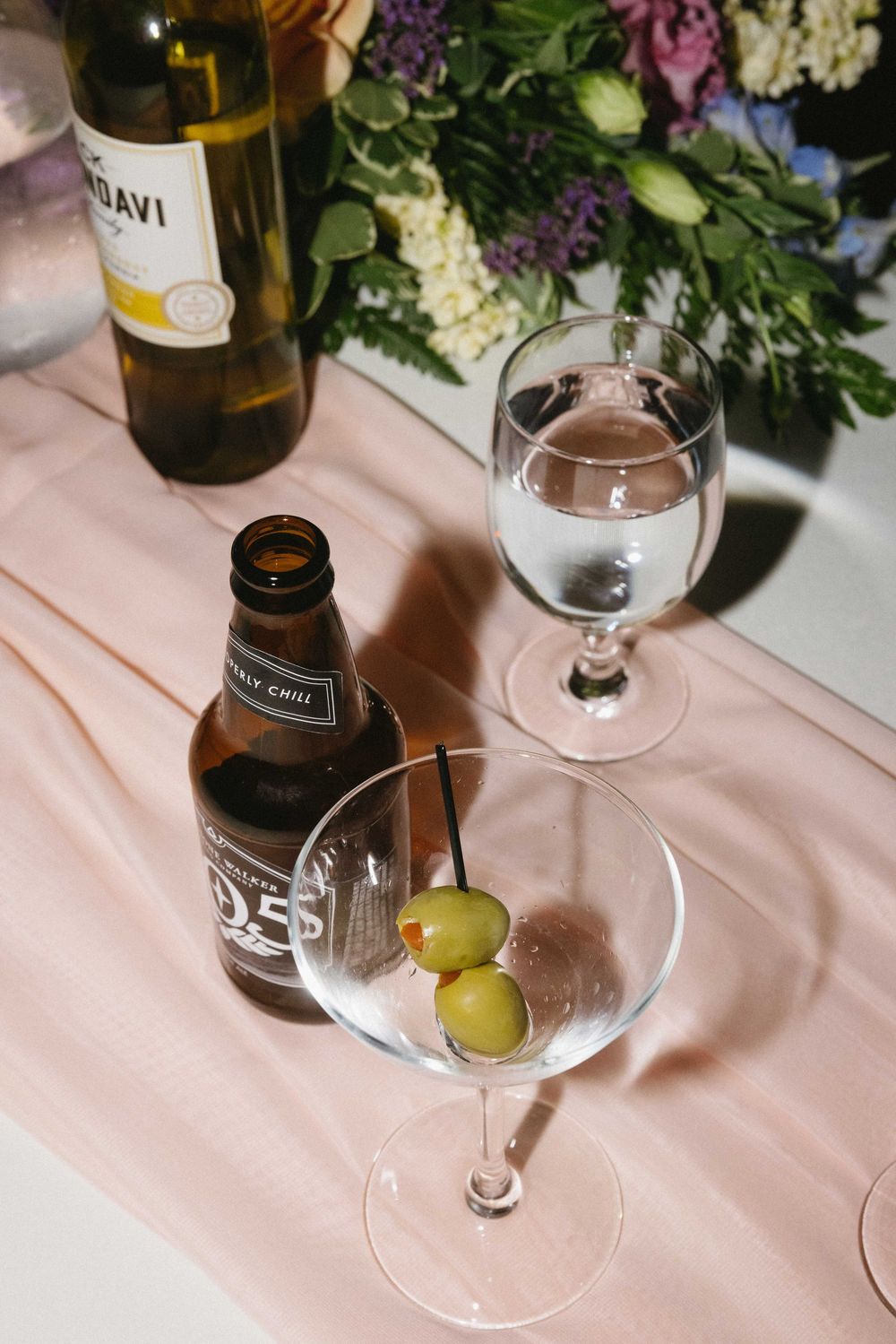 A drink setup with martini glasses, olives, and a wine bottle on a pink tablecloth next to floral arrangements.