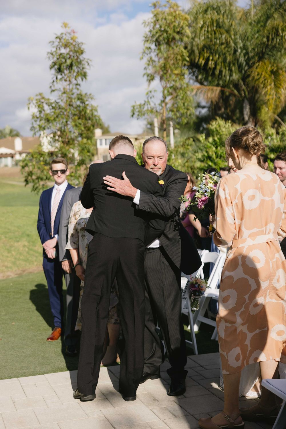 Two people embrace during an outdoor wedding ceremony while others look on near trees on a sunny day.