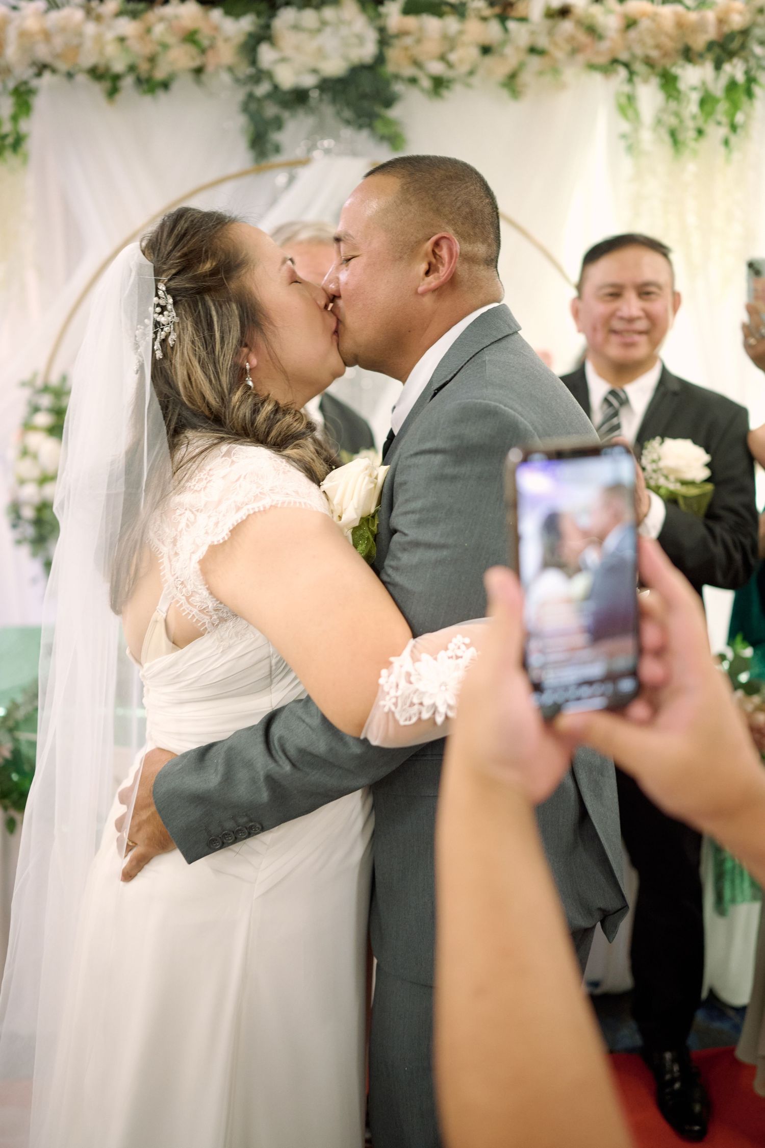 A newlywed couple shares their first kiss at the altar while someone captures the moment on a phone.