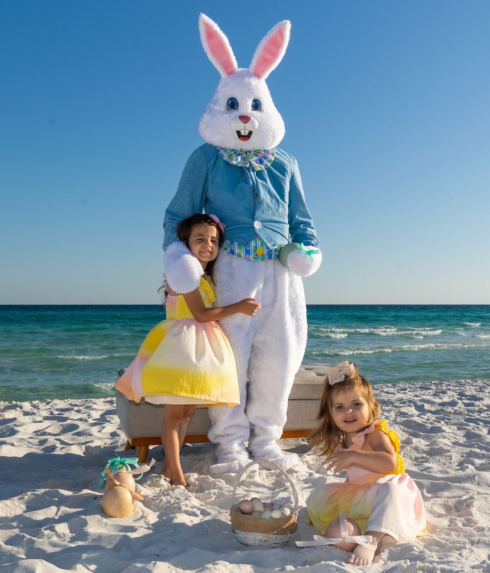 Children playing on a sunny beach with the Easter Bunny mascot near the turquoise ocean water.