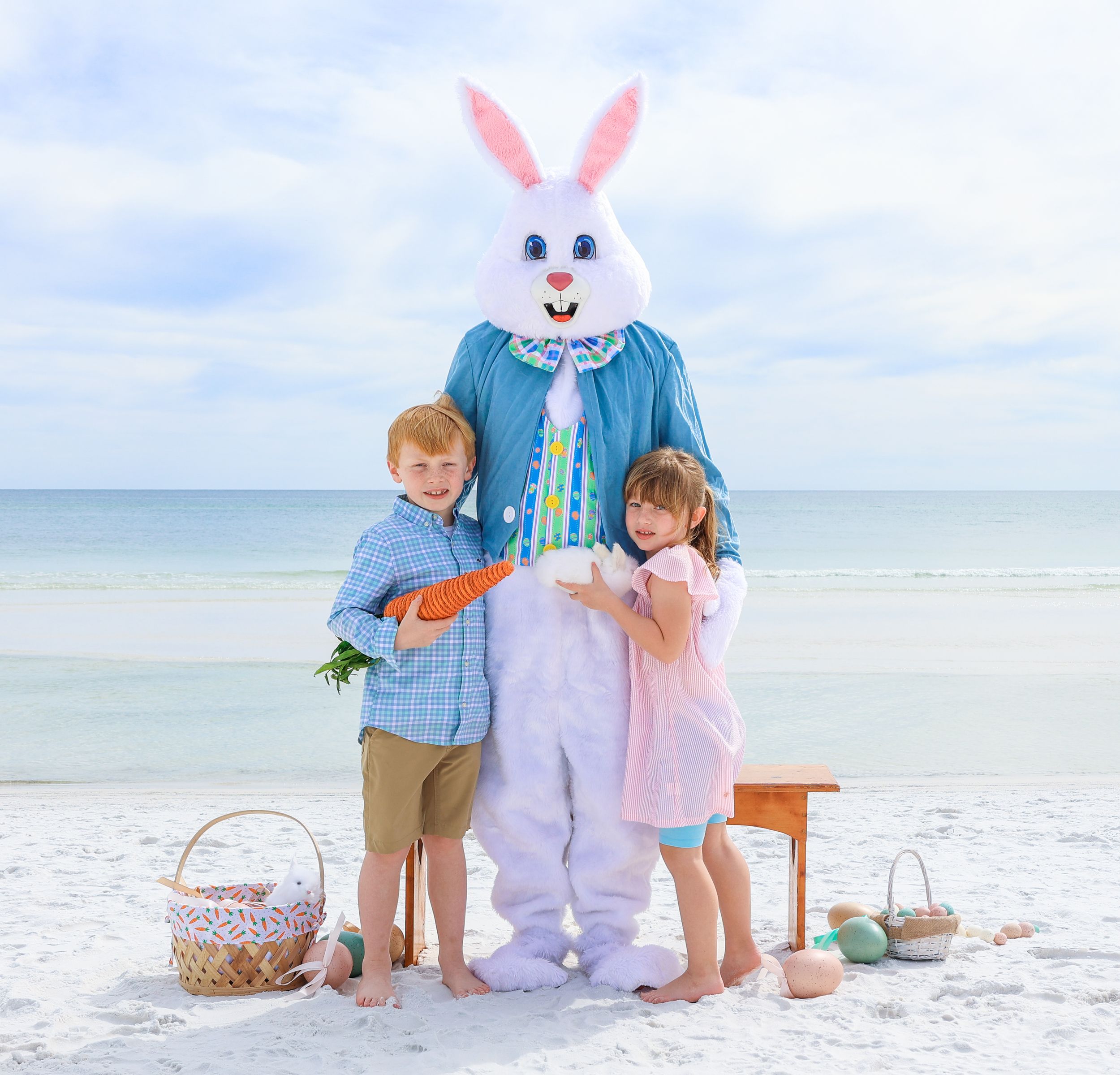 Children posing with Easter Bunny mascot on sunny beach with baskets and decorations.