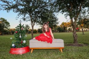 Someone in a festive red dress poses on a modern bench outdoors with a decorated Christmas tree in the background at sunset.