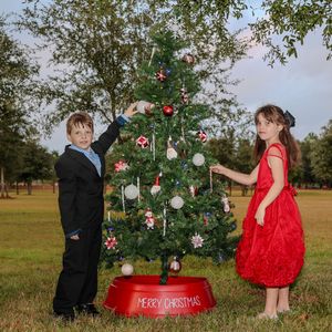 Two children decorating an outdoor Christmas tree with ornaments and decorations on a bright grassy field.