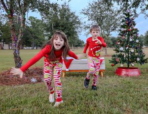 Children in matching red and white holiday outfits run playfully near a decorated outdoor Christmas tree on grass.