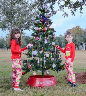 Two children in matching red and striped pajamas decorating an outdoor Christmas tree in a grassy field.