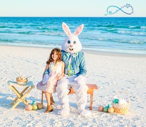Easter Bunny in colorful clothes sits with a small child on a beach surrounded by baskets and decorations.