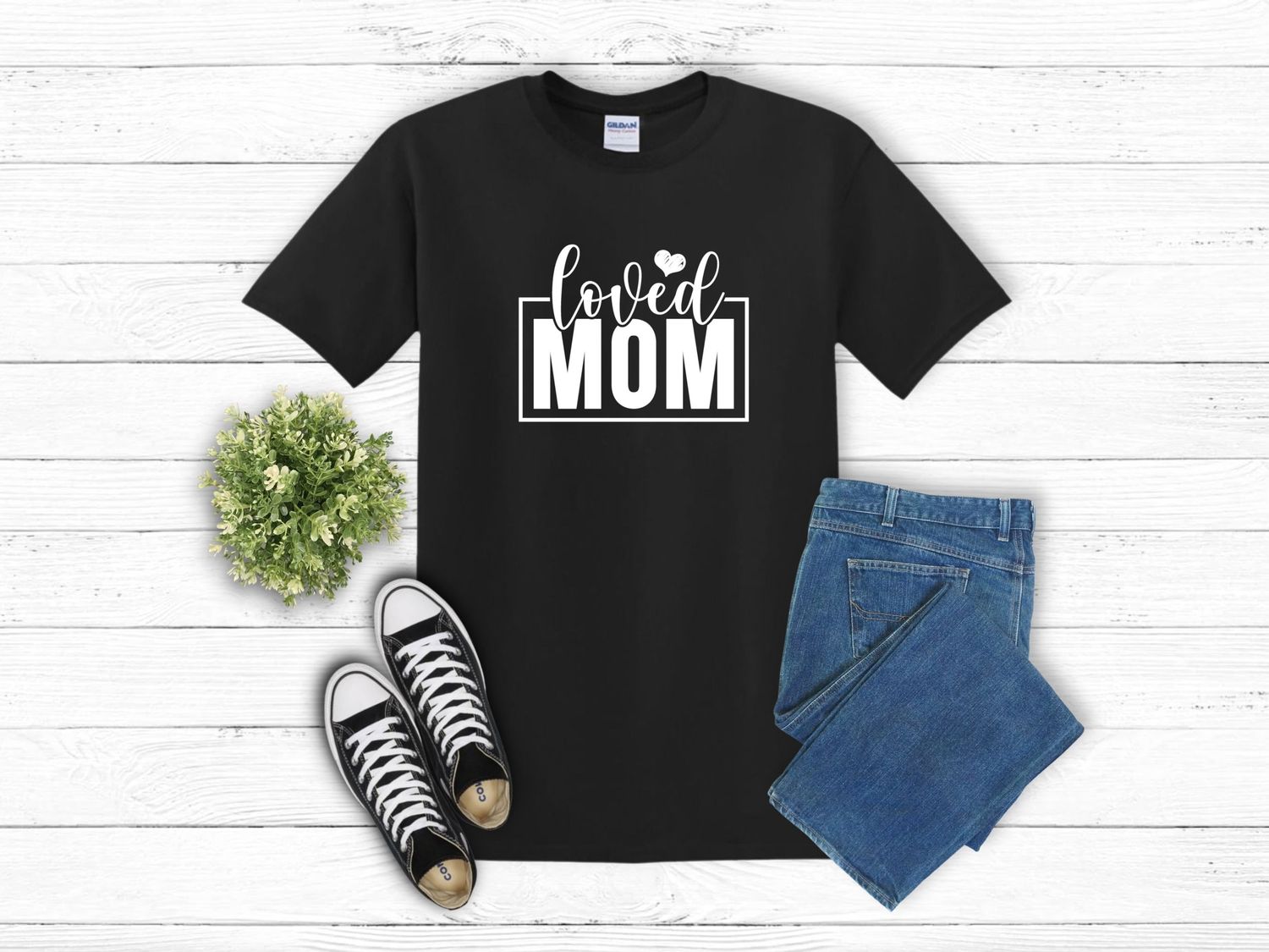 Flatlay of black baseball mom t-shirt styled with classic sneakers, blue jeans and small green potted plant on white wood.