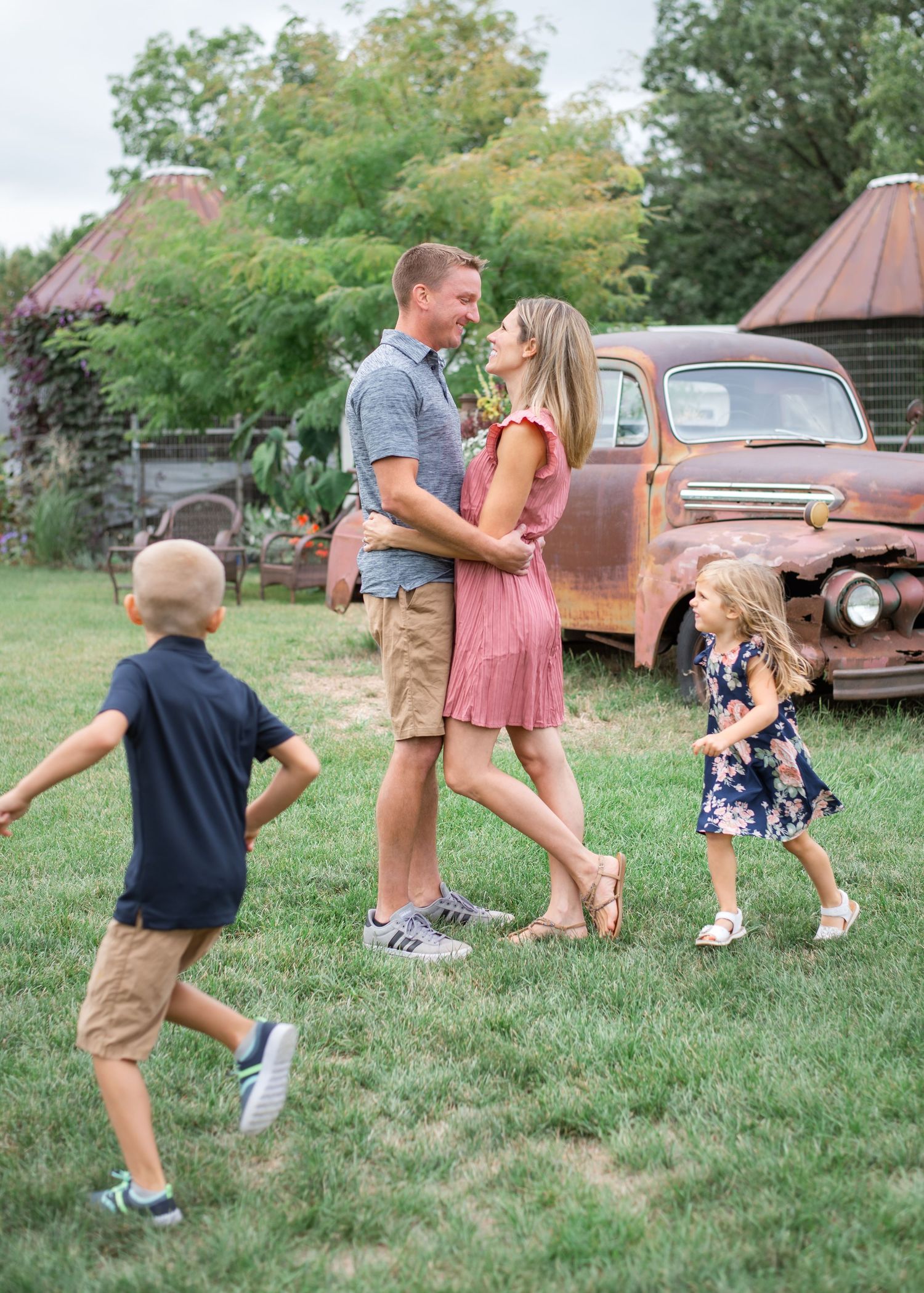 A family poses together in a rustic setting with a vintage rusty truck and barn buildings in the background.