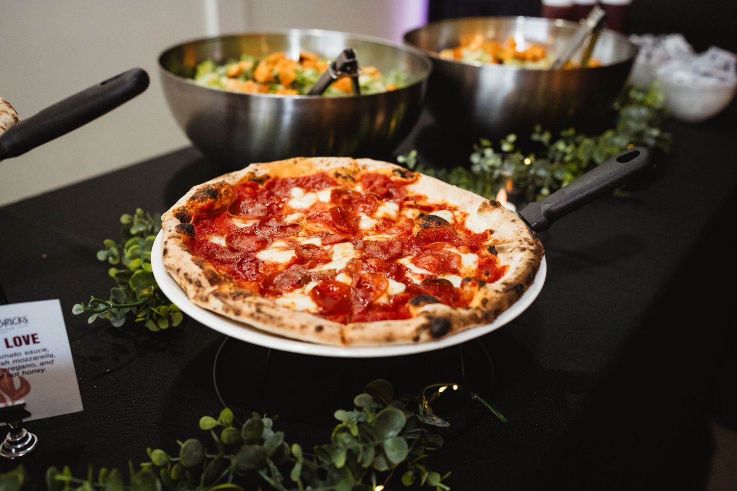 A pepperoni pizza on a white plate sits on a black table with fresh herbs and other dishes in the background.