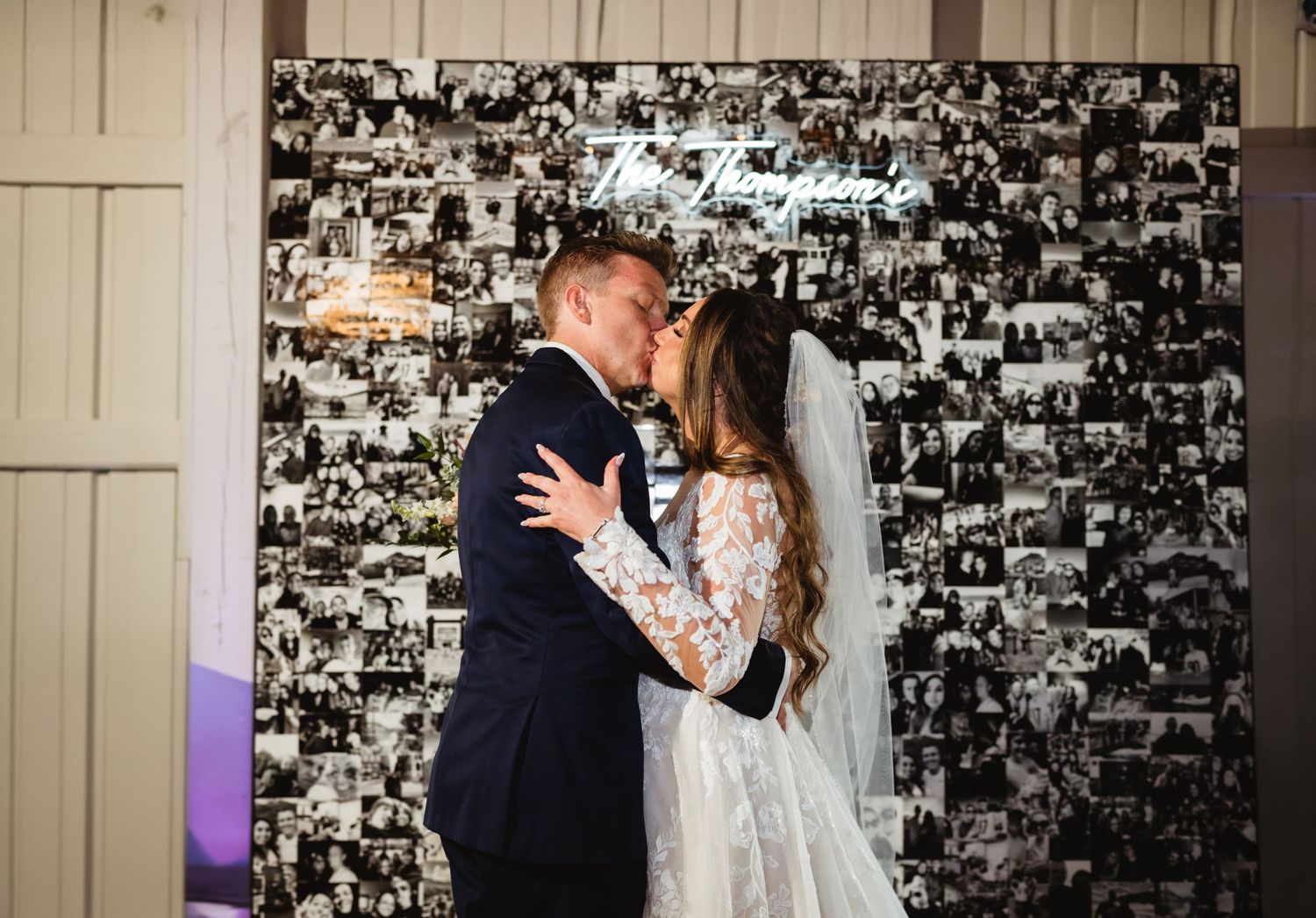 A couple shares a romantic kiss in front of a black and white photo collage wall at their wedding celebration.