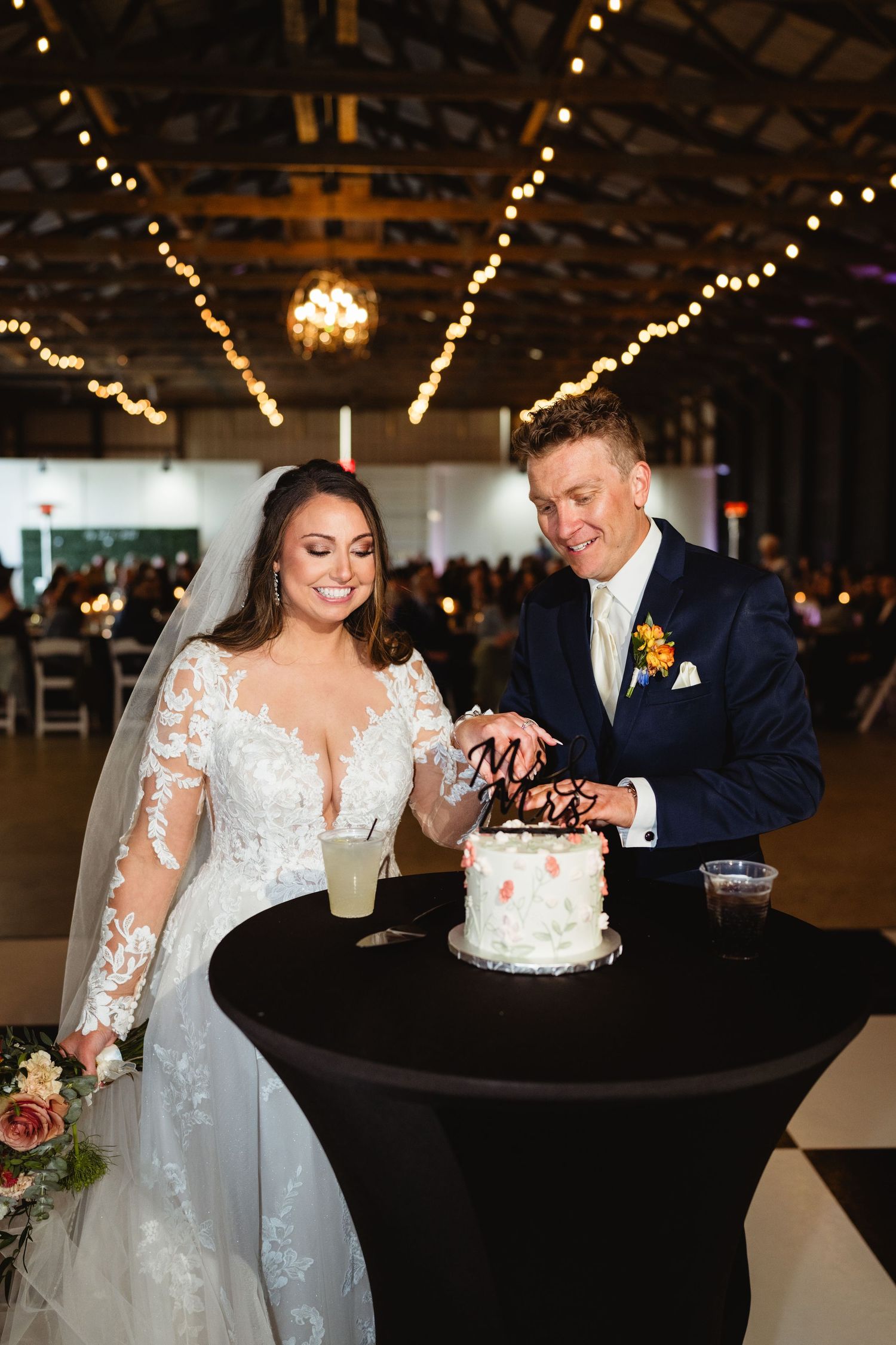 Newlyweds cut their elegant white wedding cake together in a rustic venue decorated with twinkling string lights.