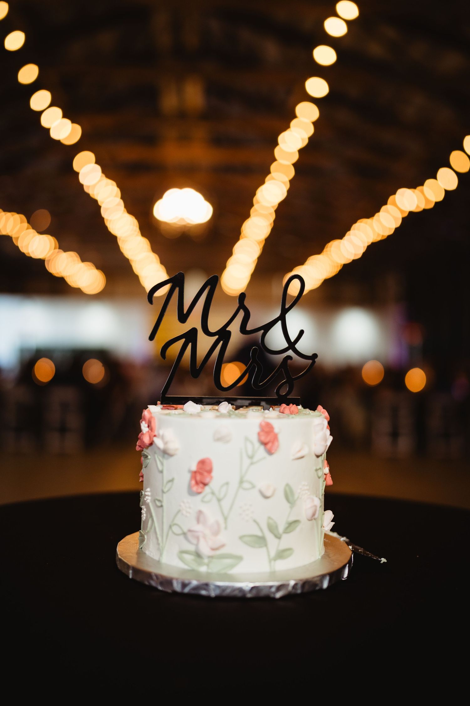 A white wedding cake with floral decorations and a Mr and Mrs topper displayed under string lights at a reception.