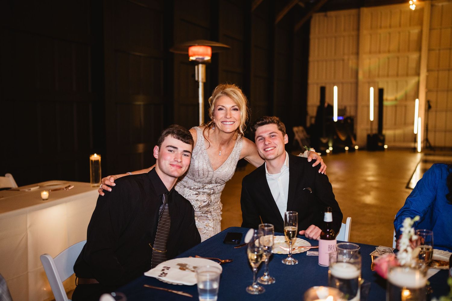 Three people in formal attire seated at a table during an elegant evening event in a spacious venue.