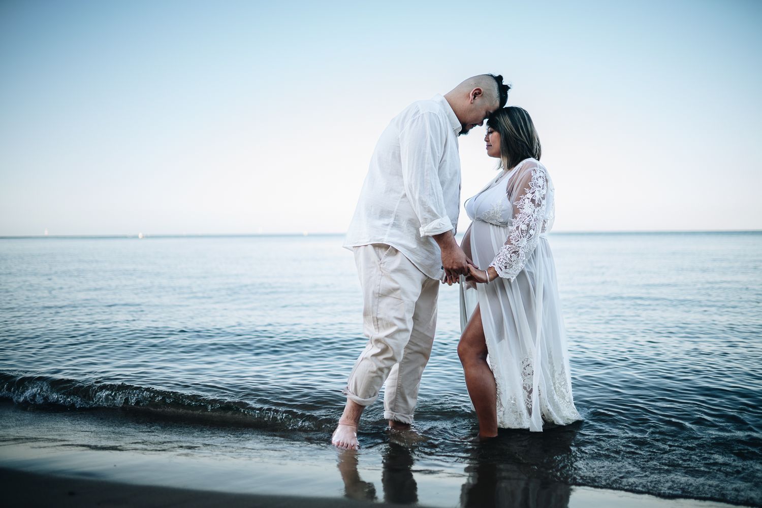Couple in a white stands facing each other with heads touching on a beach during golden hour taken by a Melbourne maternity photographer.