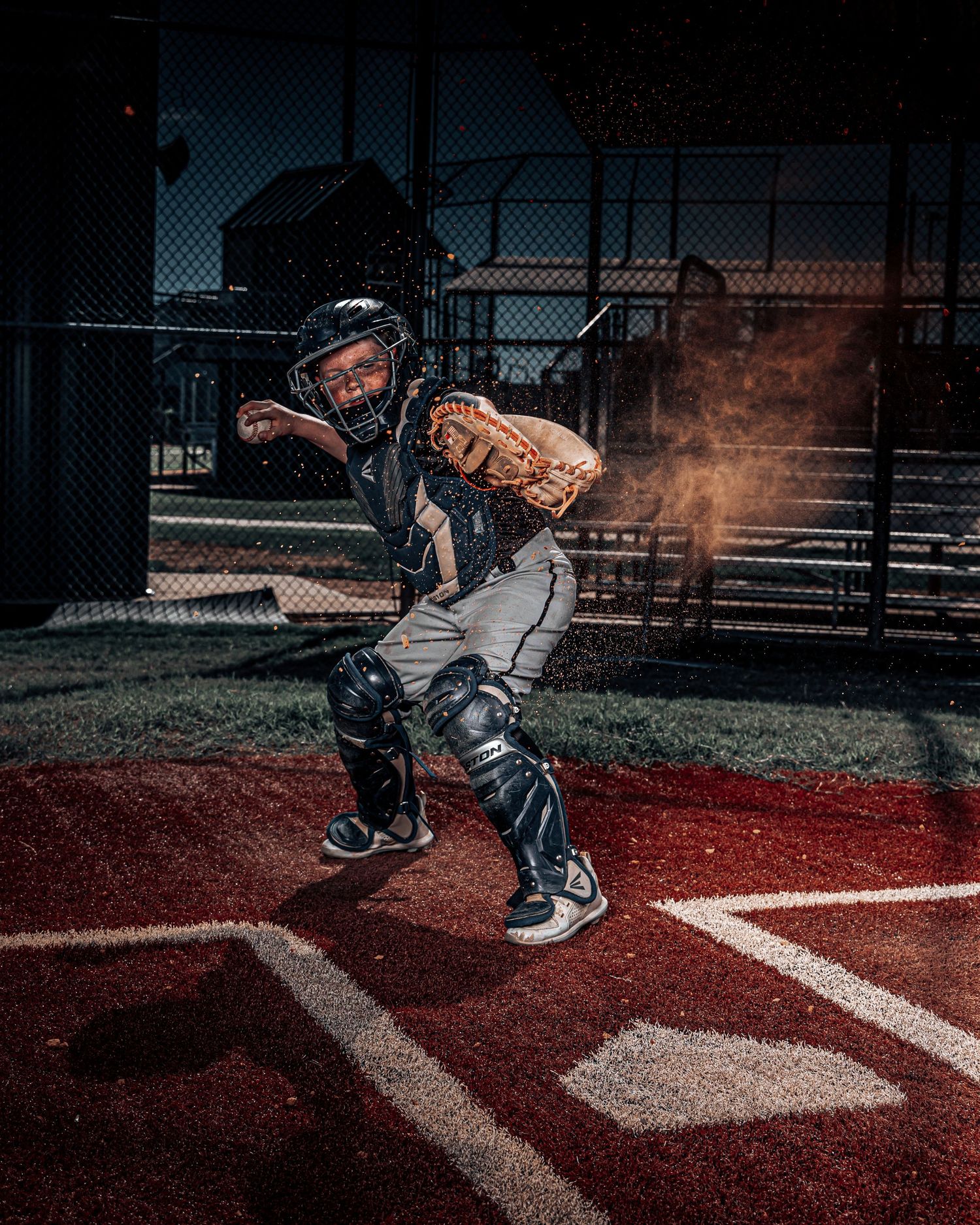 Baseball player in catching stance at home plate on red clay during nighttime practice with dramatic lighting effects.