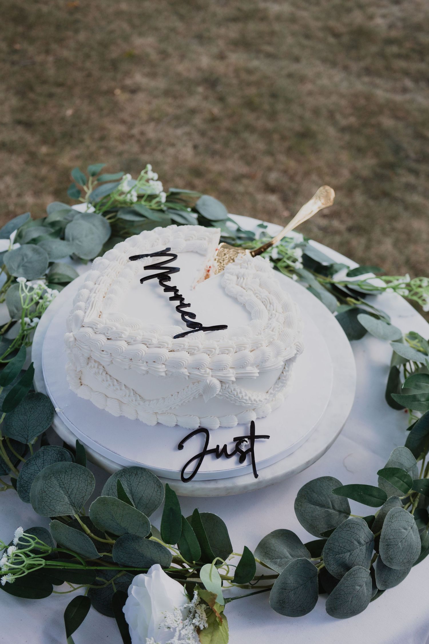 A white frosted wedding cake with decorative piping sits partially cut on a plate surrounded by green eucalyptus leaves.