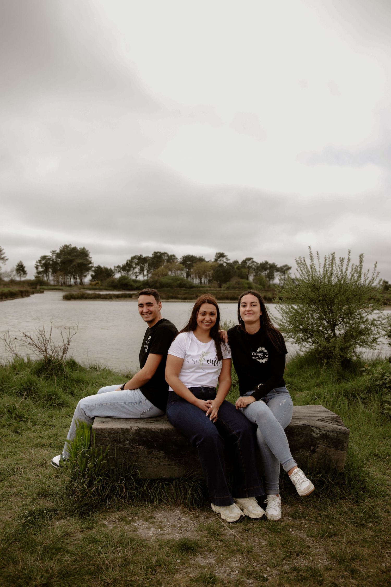 Three people sit together on a stone bench by a tranquil lake on an overcast day with trees in the background.