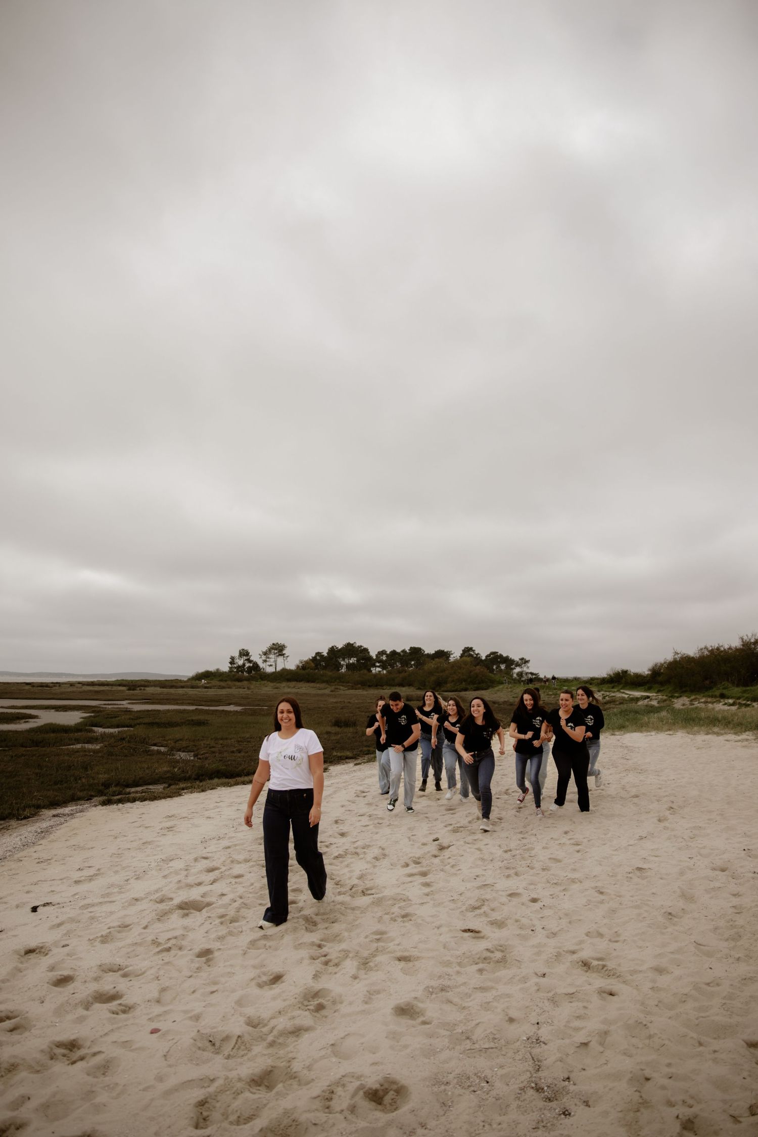 A group of people walking along a cloudy beach with marshland and trees visible in the background.