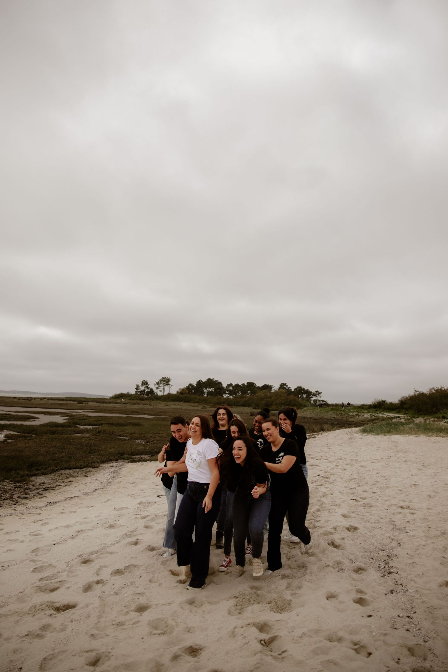 Group of people in dark clothing standing together on a sandy beach under an overcast sky.