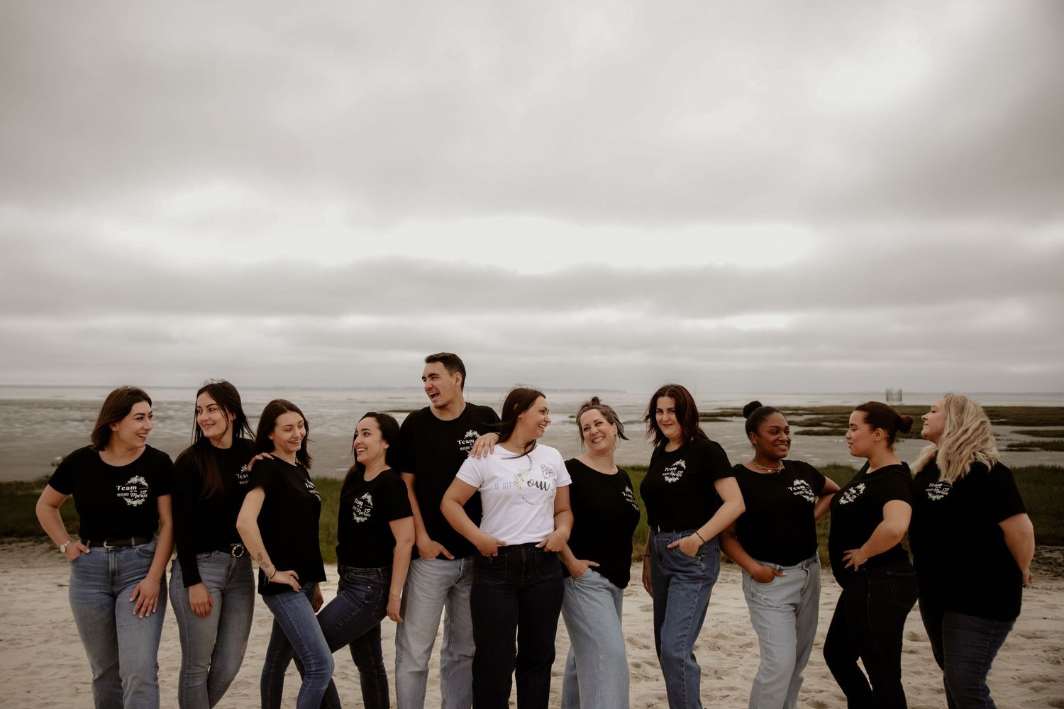 Group of friends wearing matching black and white t-shirts stand together on cloudy beach.